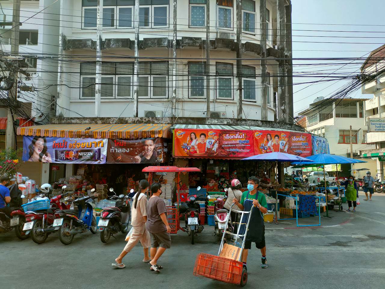 A busy street corner in Lampang, Thailand, featuring a market with colorful advertisements and people walking, surrounded by motorbikes and street vendors.