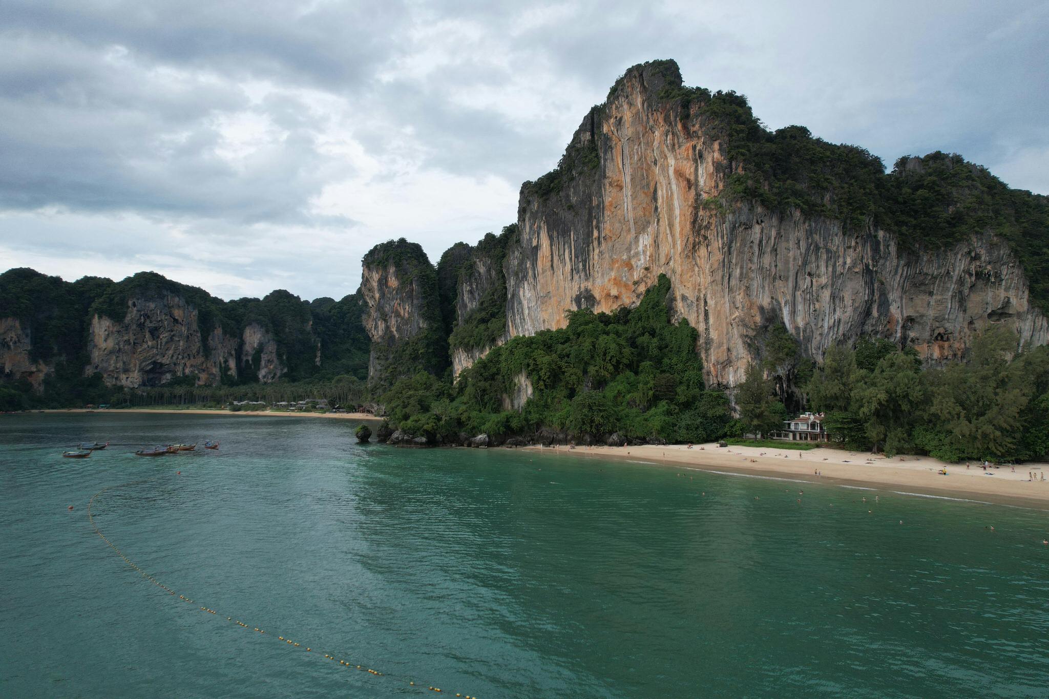 View of Railay Beach with towering limestone cliffs, lush greenery, and calm turquoise waters under a cloudy sky